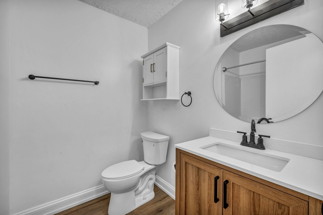 A bathroom with a white toilet, wooden vanity, and round mirror